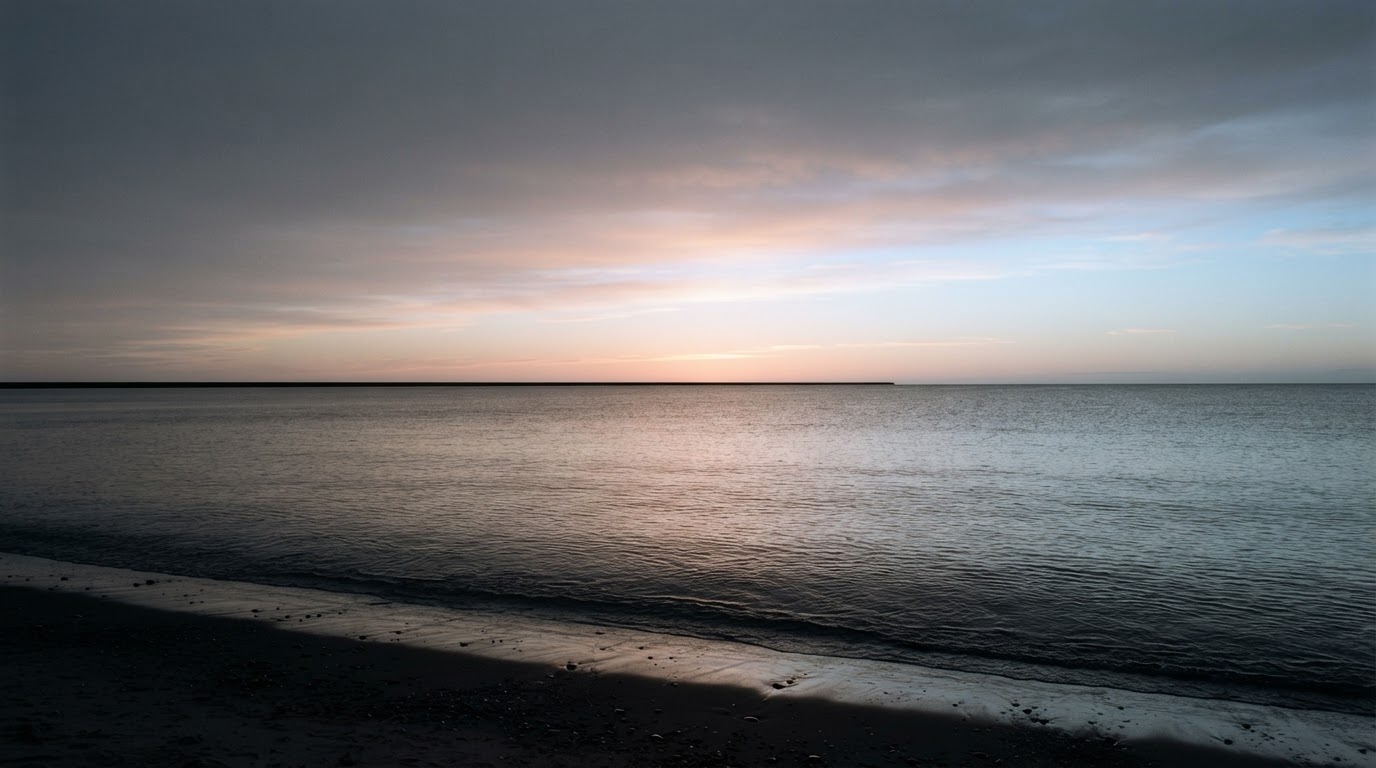The horizon of a quiet sea at dawn, the sky brightening slowly out of gray, the water even and empty (AI-generated image)