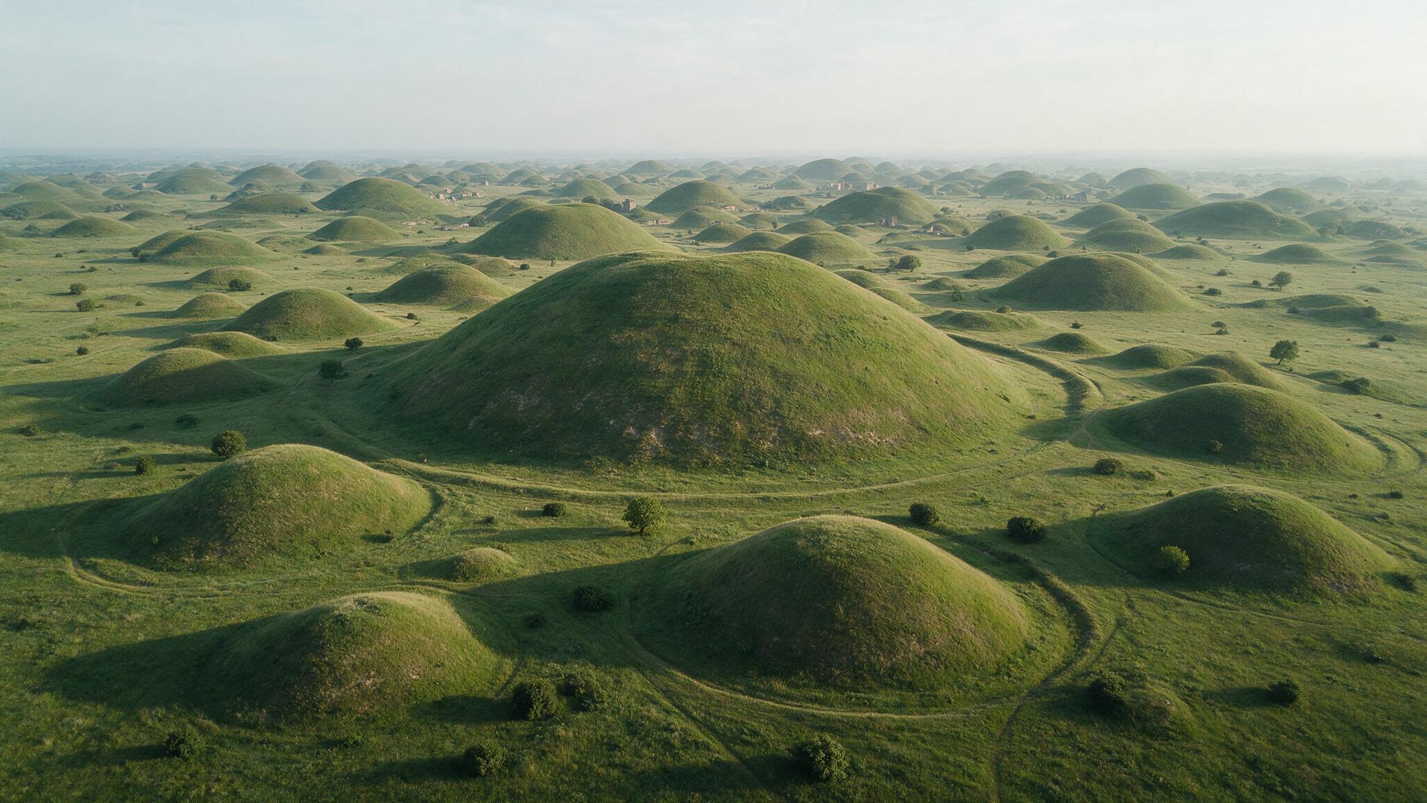 やわらかな光のもとに広がる慶州の広大な芝の古墳群を、上から見下ろすような光景（AI生成画像）