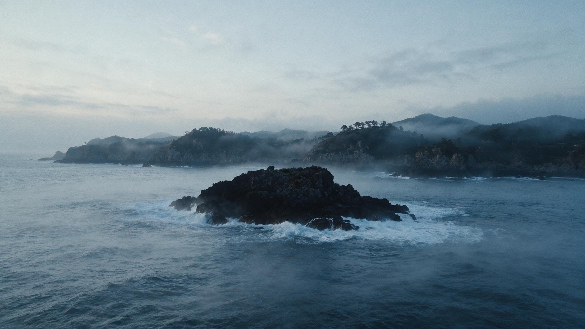 A distant view of a lone low rocky islet standing amid the crashing waves of the East Sea, dawn mist, an air of mystery (AI-generated image)