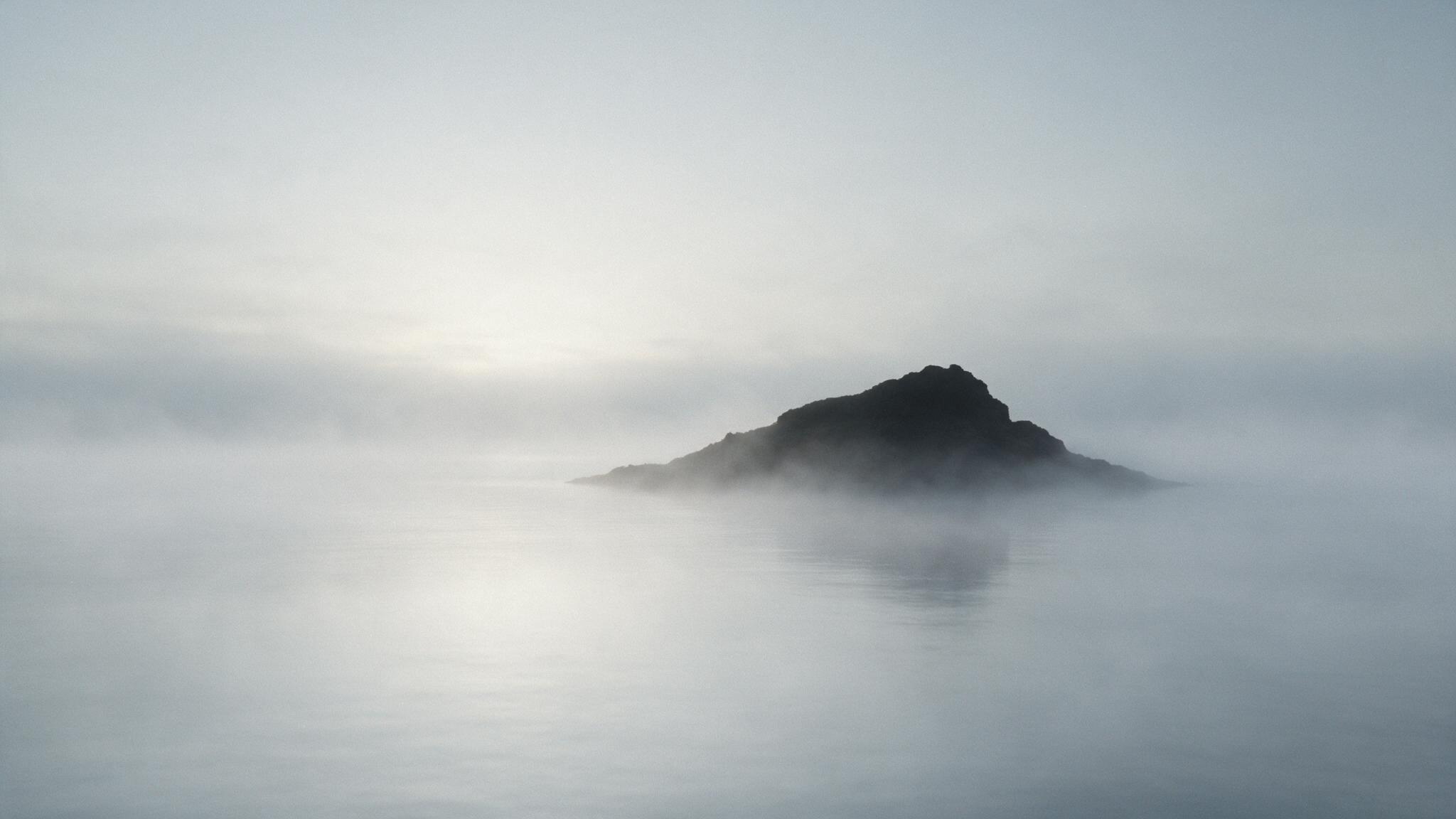 A rocky islet rising in silhouette over a fog-shrouded sea at dawn, a still and mysterious mood (AI-generated image)