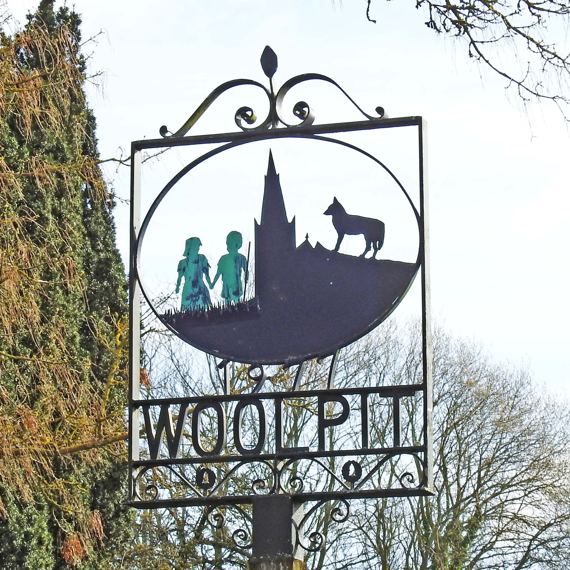 A close view of the Woolpit village sign: two green children holding hands, set beside the church spire and the wolf on the village emblem