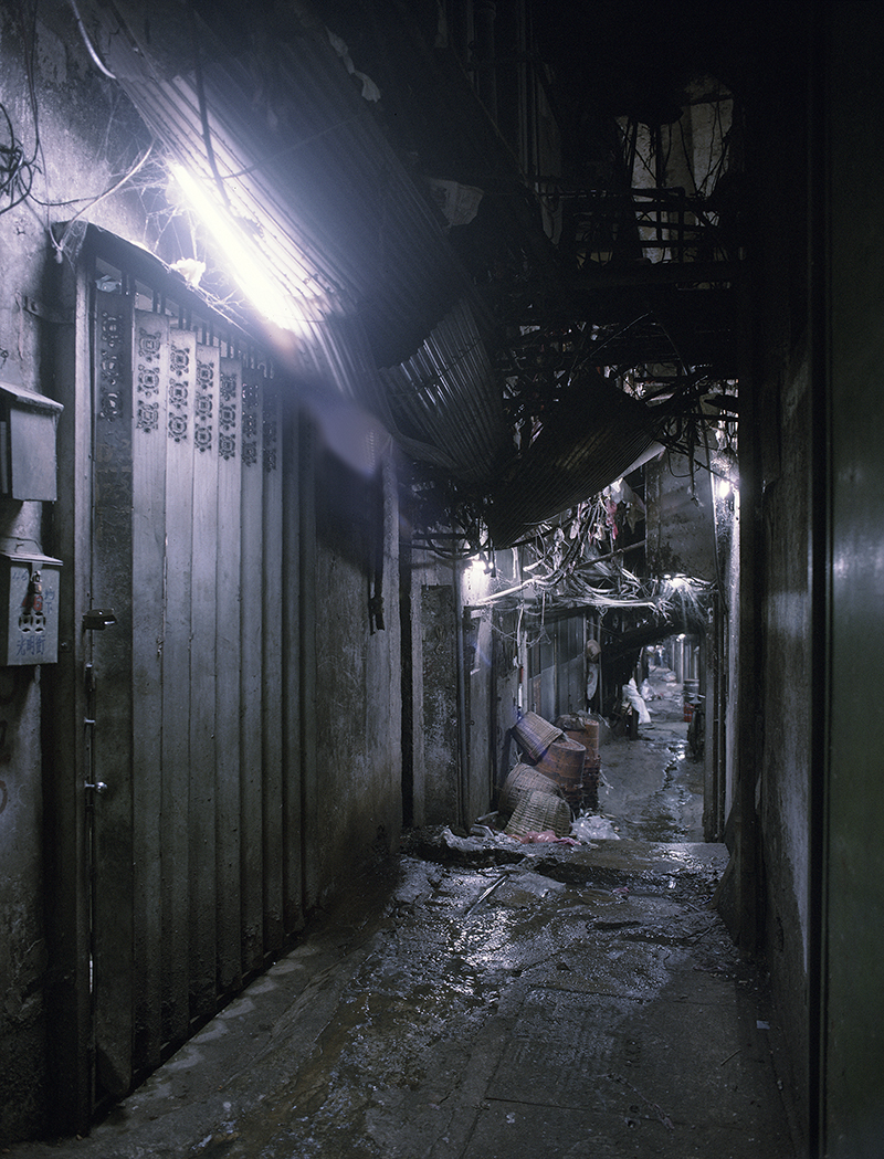 A dark alley inside the Walled City — a real passage with a wet floor, drooping cables, and a single fluorescent tube.