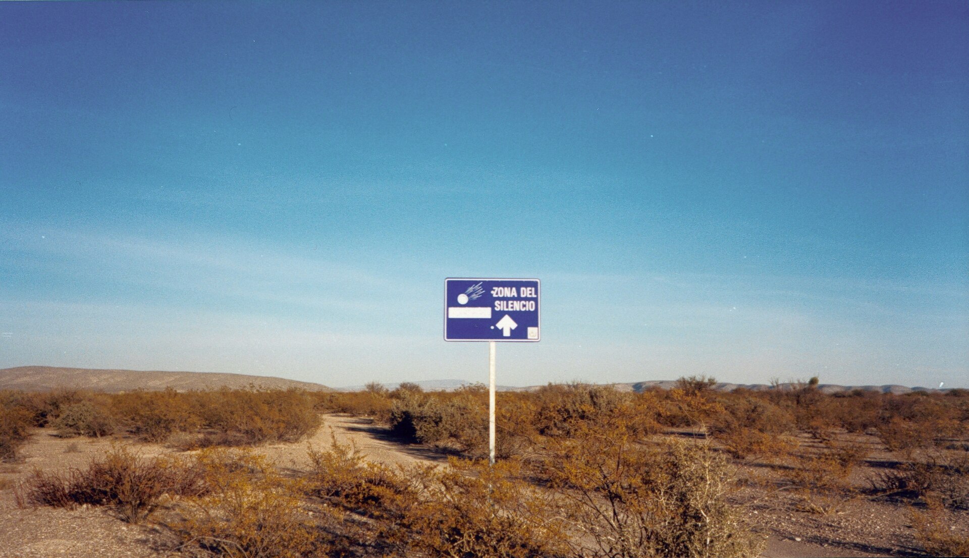 A lone "Zona del Silencio" road sign, marked with a falling meteor, standing among dry scrub in the middle of the desert
