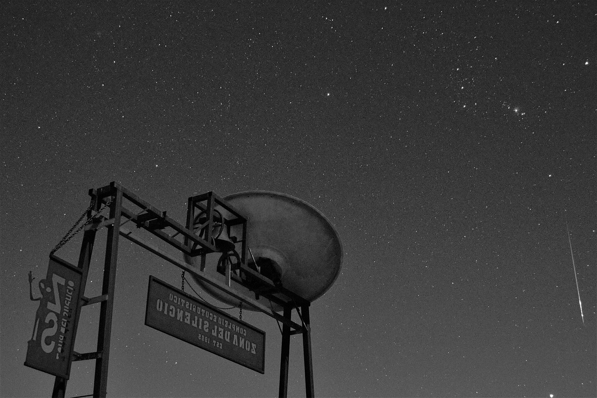 The parabolic dish and signboard of the Zona del Silencio observation site seen from below against a sky thick with stars, a meteor streaking down at right